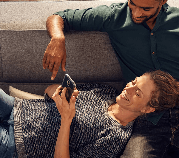 Smartfon z aplikacją Home Connect z dodatkowymi programami do zmywarki. W tle kobieta i mężczyzna na kanapie.